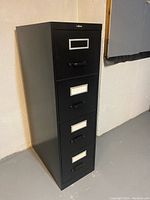 Photo of the black 4-drawer metal file cabinet showing front and side, metal frame label holders on drawers, black drawer handles, vertical design.