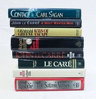 Stack of 8 hardcover books showing titles and authors on spines: 'Contact' by Carl Sagan, 'A Most Wanted Man' by John Le Carré, 'Ways of Escape' by Graham Greene, 'The Bourne Supremacy' by Robert Ludlum, 'The Mission Song' and 'Smiley's People' by John Le Carré, 'Lie Down With Lions' by Ken Follett, and 'The Satanic Verses' by Salman Rushdie.
