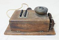 Side angled view showing wooden box, metal ringer bell, push buttons in two rows, and earpiece wire.