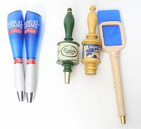 Five beer tap handles arranged on white background: two blue and white Sam Adams Boston Lager handles, green Carlsberg, yellow Great Lakes, and beige Sam Adams with blue plexiglass insert