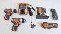 Photo showing two orange and black Rigid 12V cordless drill drivers, a charger with power cord, jigsaw attachment head, impact attachment head, and a base tool handle/body laid out on white surface.