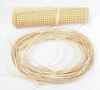 Two rolls of cane for chair repair. One roll of narrow thin cane strips coiled on the floor beside a roll of woven cane mat with open weave pattern tied with a beige strip.