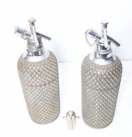Two glass seltzer bottles wrapped in metal mesh and chrome siphon tops standing side by side with a small metal CO2 holder between them.