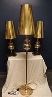 Full view of the 3 lamps against dark blue background on white covered table, two shorter table lamps with brass bases and one tall brass floor lamp, all with gold mylar shades.