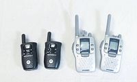 Photo showing both pairs of walkie talkies side by side on a white background. Two larger silver Sylvania radios on the right, two smaller black Motorola radios on the left.