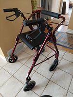 Front and side view of a red aluminum collapsible walker with black padded seat, arm rests, and brake lock handles shown on tile flooring.
