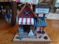 Front-facing view of the custom birdhouse with detailed workshop facade, awning, tools, signage, and tricycle figurine on base.