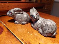 Two gray hand made cultured stone rabbit figurines with red eyes on wooden surface, showing detail and size difference.