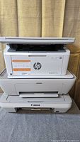 Stacked view of three printers: top HP Laserjet M139we, middle HP Deskjet 2755e, bottom Canon MG2420 printer.