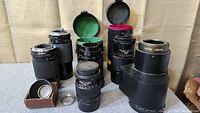Six vintage 35mm camera lenses including a large Letus Nikon adapter and three filters, arranged on fabric surface with some lens caps and cases.