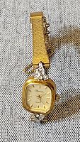 Gold-tone Rochelle ladies watch with rectangular face and rhinestone decorations, on a textured light grey fabric background.