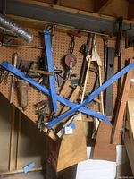 Wide view of antique wooden and metal hand tools hanging on pegboard wall including saws, hand planes, clamps, wooden trowels, pulley wheels, and other woodworking tools.