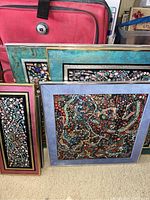 Photo showing five framed pictures featuring bead art leaning against a red suitcase on the floor.