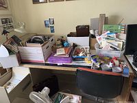 Wide view of table holding multiple boxes and containers filled with art and drafting supplies, including papers, folders, stamps, crafting tools, and various small items. Portable lamp and fan positioned nearby. Chair in front of table and briefcase under table visible.