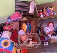 Photo of assorted vintage dolls, including Raggedy Ann and Ken, doll clothes stored in a wooden closet and plastic bin, a small mirror, and a crocheted hat accessory on a couch.