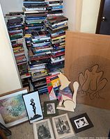 Stacked hardcover and softcover books with visible spines and titles, two atlases at front including Wyoming Atlas, surrounded by several framed and unframed prints and art pieces, wooden dog figurine with scarf, and wood coyote silhouette wrapped with scarf