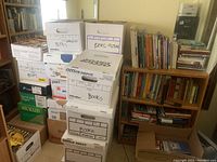 Photo showing stacked boxes labeled 'Books' containing various books next to a small wooden shelving unit filled with books.