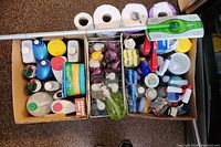 Top view of three cardboard boxes containing a variety of cleaning products arranged tightly. Visible items include paper towels, several bottles of liquids, sponges, hand soaps, Lysol wipes, and the Swiffer mop handle and pads.