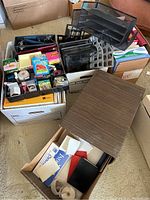 Boxes containing assorted office and teacher supplies including binders, stationery, envelopes, Post-it notes, tape, notebooks, and desk organizers.