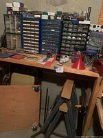Wide view showing multiple plastic storage organizers on a wooden table, holding various fasteners and small hardware. Tools and accessories scattered on surface along with a dolly and two folding chairs below the table.