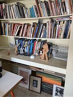 View of shelves with numerous books and several handmade sculptures displayed on a shelf under the books. Wire shelves are also present.