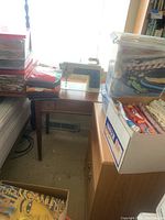 Wide view showing antique Singer sewing machine set in a wooden table near a window, surrounded by multiple bins and boxes of fabric on and next to the table.