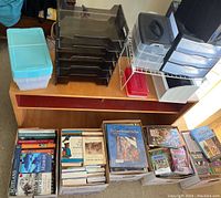 Wooden shelf unit with various office storage bins, black tiered trays, plastic drawer organizers and boxes of books beneath