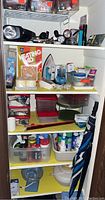 Four shelf closet containing various utility items including flashlights, a vintage GE heating pad box, a Sunbeam iron, multiple household cleaning and utility supplies, and umbrellas.