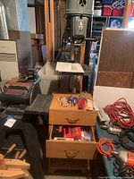 Image showing Rockwell band saw next to wooden two drawer cabinet filled with various blades and bits. Additional tools like hand drills and cords also visible.