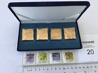 Open blue velvet box holding five gold-plated square medallions; below, four Canadian stamps and ruler