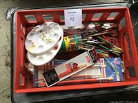 Top view of a red plastic crate containing artist paintbrushes, plastic and ceramic palettes, and various art drawing tools.