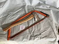 Photo of a hand axe and a metal buck saw with orange frame laid out on a white cloth background.