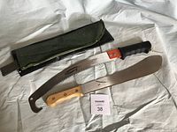 Photo shows two outdoor cutting tools: a curved serrated bush saw with black handle and green fabric sheath, and a metal machete with wooden handle.