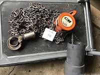 CM chain hoist with large chain coiled beside it, metal hook attached at the end of the chain, photographed on a black textured surface.