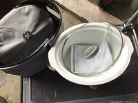 GE slow cooker crock pot with glass lid and olive branch pattern on the body, placed next to a black insulated carrying bag with GE logo.