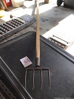 Photo of the fishing spear laid out showing full length, wooden handle, and metal five-tine spear head, slightly rusted.