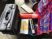 Black leather expandable lawyer briefcase, red and blue organizer, multi-outlet power strip with cords, and rolled-up red air mattress arranged on floor.