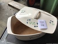 Ceramic lid of the roasting pan is cream colored with blue and green floral patterns, shown resting on top of the cast iron pan.