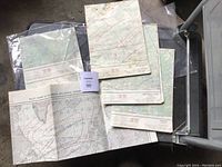 Five vintage maps displayed on a black surface, including a nautical pilot chart and regional topographic maps, some protected in clear plastic sleeves.