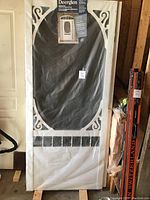 Full view of new Deerglen composite Victorian screen door wrapped in protective packaging, showing white frame with decorative corner trim and black screen insert.