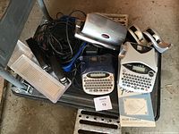 Two Brother P Touch label makers (one gray, one blue) with keyboards and small LCD screens, a metal stapler, two tape dispensers, and assorted electronic cables on a black tray on the floor.