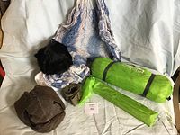 Photo showing a green tent bag, a smaller green bag with poles or pegs, a blue and white hammock, and two furry winter hats (brown and black) on a neutral cloth background.
