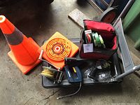 Overview photo showing two orange collapsible safety cones, a hand winch with yellow rope, a red bag with rolls of painters tape, and an electric staple gun on a black cart.