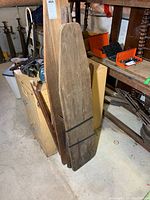 Two antique wooden ironing boards leaning vertically against a filing cabinet and nearby furniture. One has black metal straps wrapped around it; surfaces show wear and scratches.