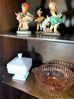 Porcelain figurines arranged on a wooden shelf with a white hobnail milk glass covered dish and a pink ribbed glass bowl below them.