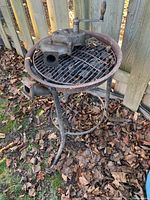 Full view of three-legged metal forge with grill top and hand-crank blower placed outdoors on grass and leaves.