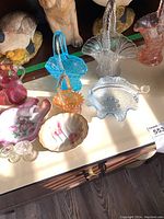 Photo showing blue, peach, pink, and clear glass baskets alongside decorated porcelain trinket dishes and small clear glass eye washers.