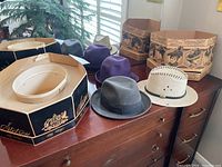 Three hats including gray Stetson Premier 1/8, purple felt hat, beige woven hat, and two vintage Stetson hat boxes on wooden surface
