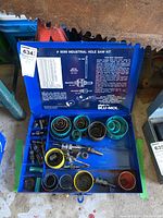 Open blue metal case showing assortment of hole saw blades, mandrels, adapters, and pilot drill bits organized inside compartments.