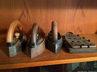 Front view of 3 vintage charcoal irons with wood handles and metal holder on a wooden shelf